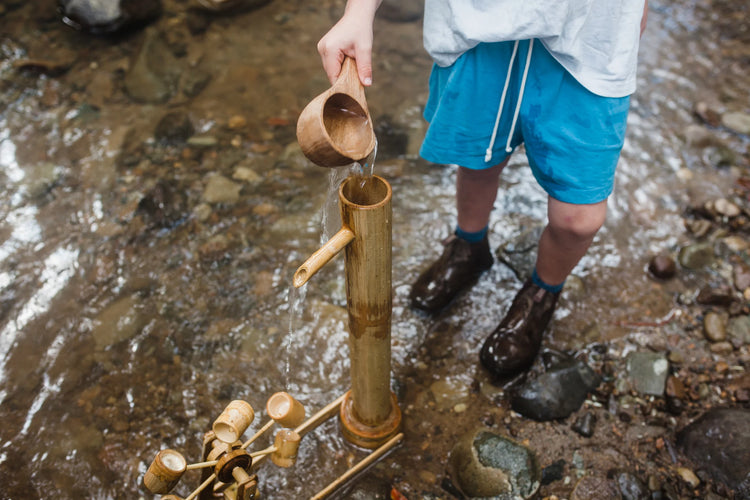 Person using a wooden cup to fetch water from a bamboo pump in a stream.
